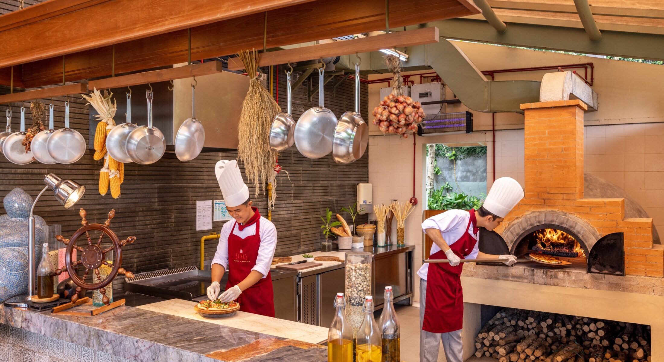 Two chefs in white uniforms and red aprons preparing fresh pizza in an open-concept kitchen next to a traditional brick oven at SereS Springs Resort's Bell'Aria Italian Restaurant